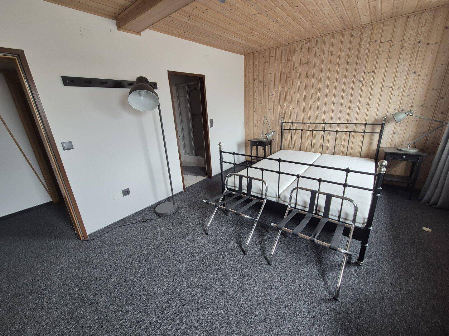 Schlafzimmer mit Holzwandverkleidung, Metallbett und grauem Teppichboden, mit Zugang zu weiteren Räumen.