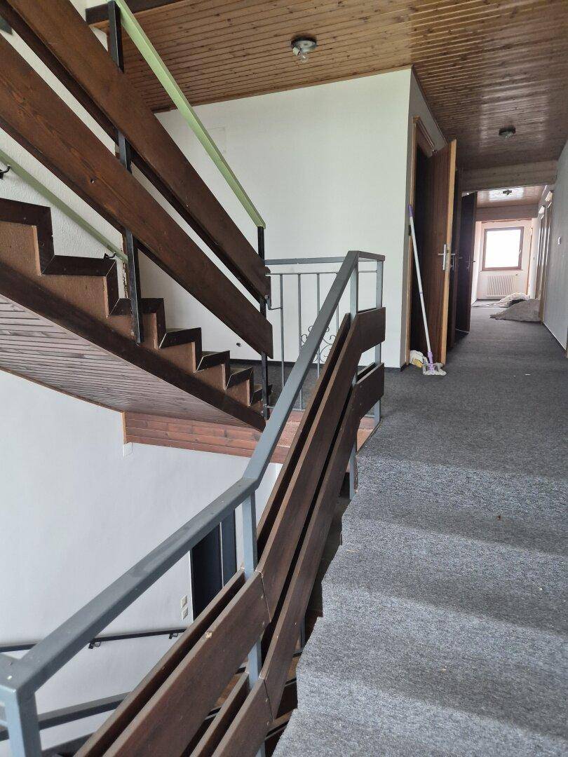 Treppenhaus mit Holzstufen und Geländer, das zu einem Flur mit Teppichboden führt.