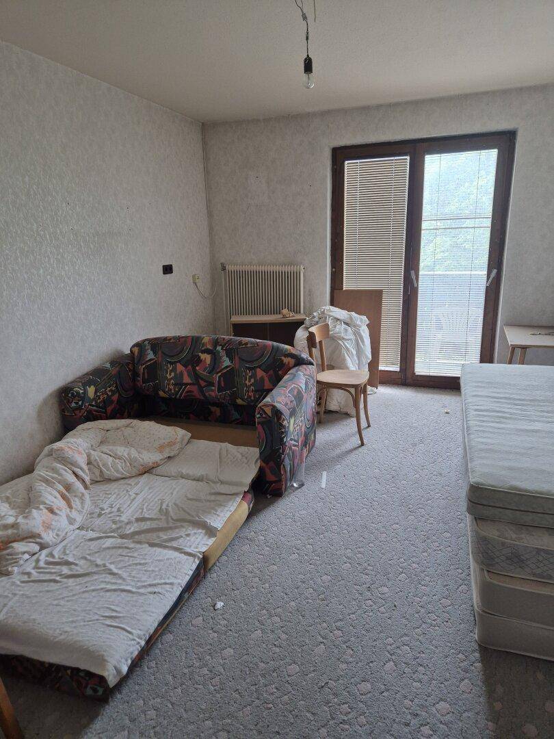Einfaches Schlafzimmer mit Schlafsofa, Matratze und großer Fenstertür mit Jalousien.