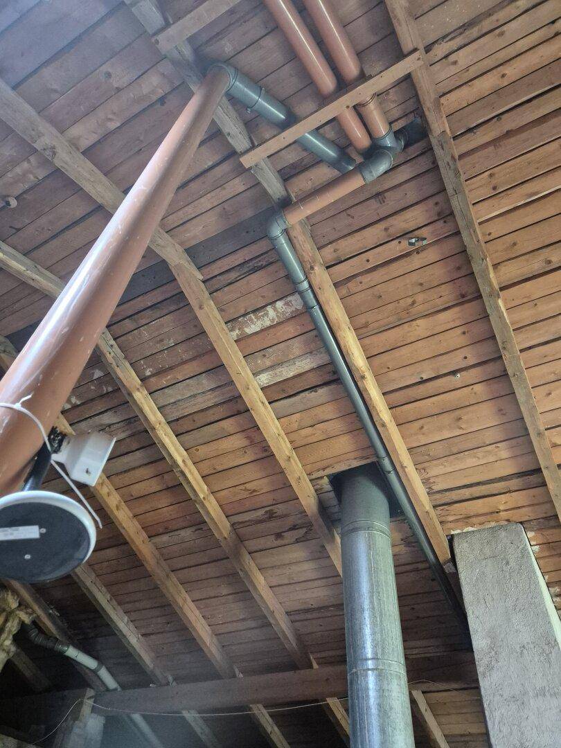 Freiliegende Holzdeckenbalken mit sichtbaren Rohren und Leitungen in einem Dachbodenbereich.