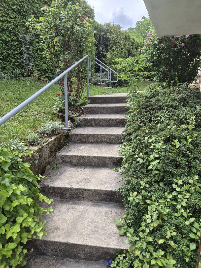 Betontreppe mit Metallgeländer, umgeben von üppigem Grün und Sträuchern in einem gepflegten Gartenbereich.