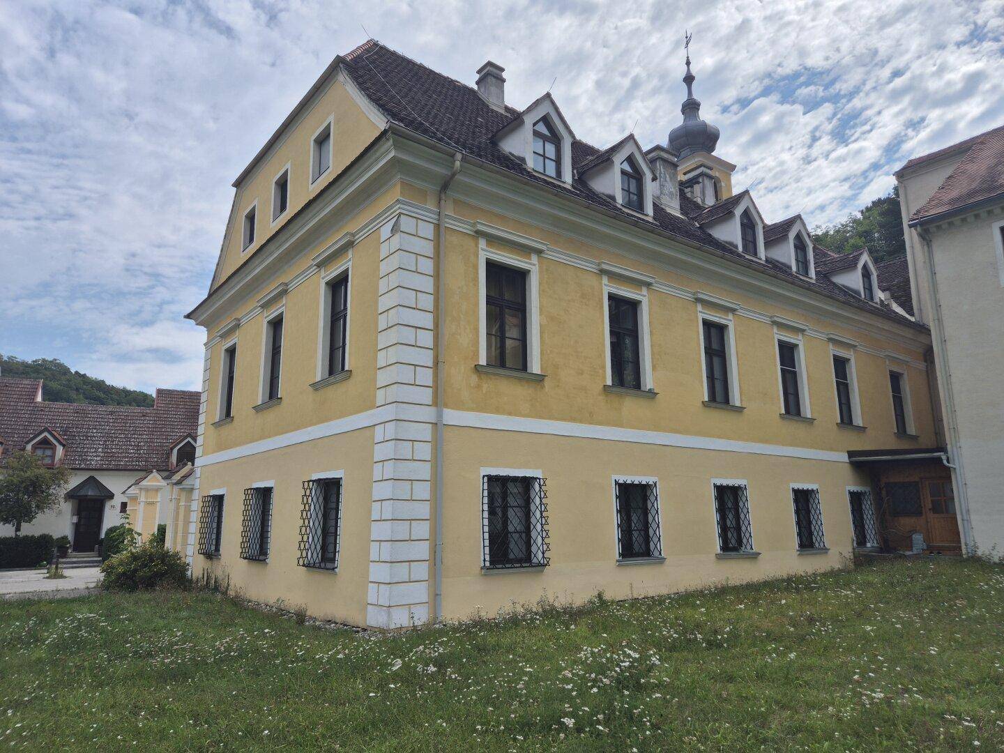 Historisches Gebäude mit gelber Fassade, Mansarddach und schmiedeeisernen Fenstergittern, umgeben von Grünflächen.