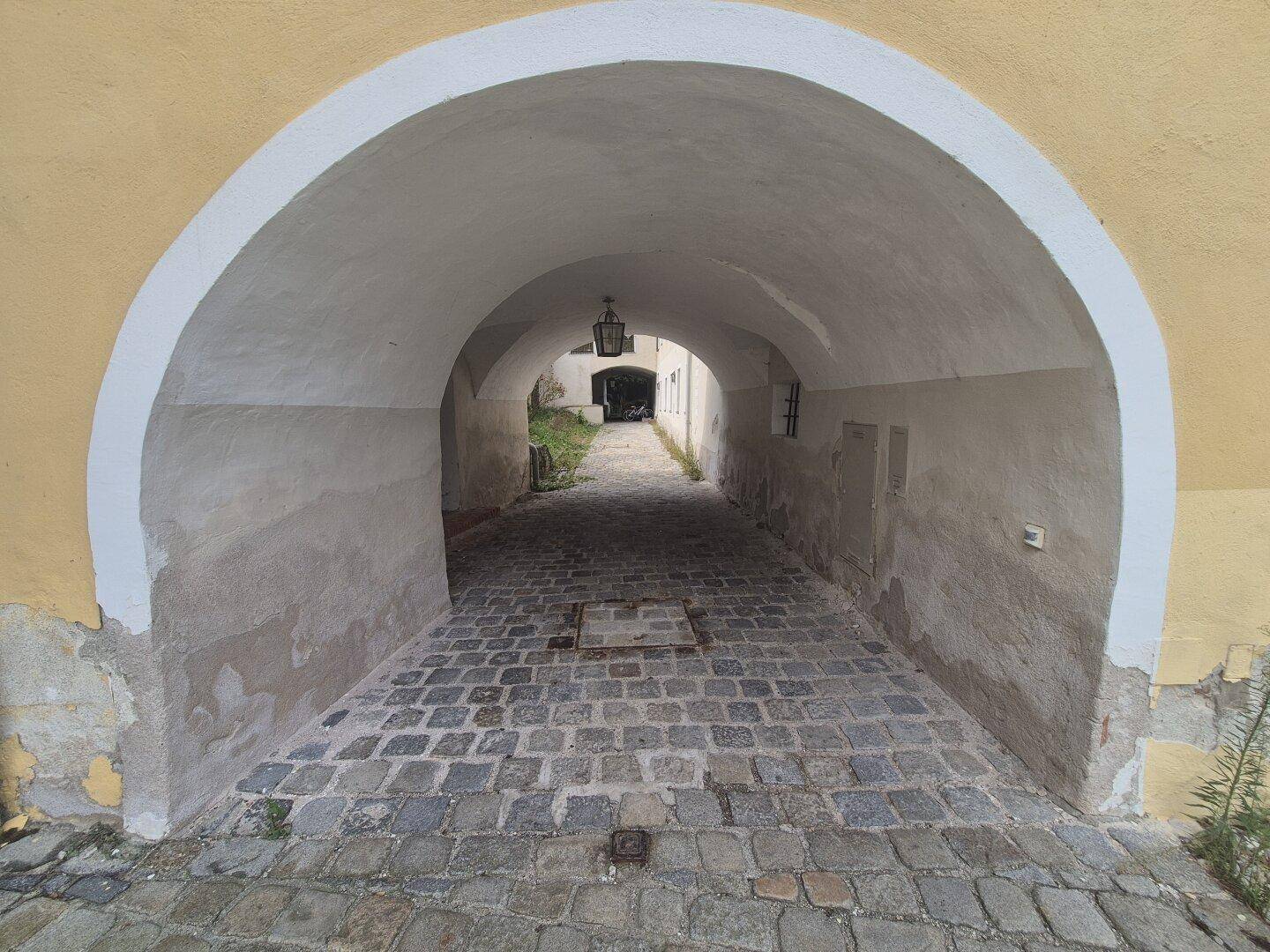 Historischer Torbogen mit Kopfsteinpflasterweg führt durch das Gebäude, beleuchtet von einer Laterne.