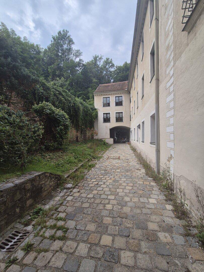 Kopfsteinpflasterweg zwischen Gebäuden und einem bewachsenen Hang, der zu weiteren Teilen des Anwesens führt.