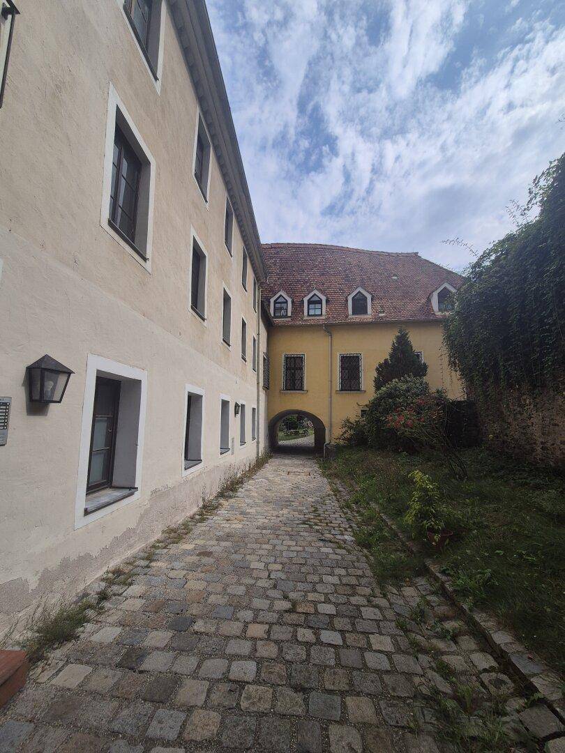 Historischer Kopfsteinpflasterweg zwischen zwei Gebäudeteilen, mit Blick auf eine gelbe Fassade und bewachsenen Hang.