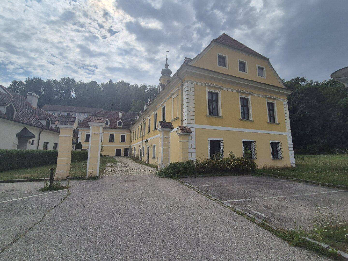 Historisches Anwesen mit gelber Fassade, Mansarddach und gepflastertem Vorplatz, umgeben von Natur.