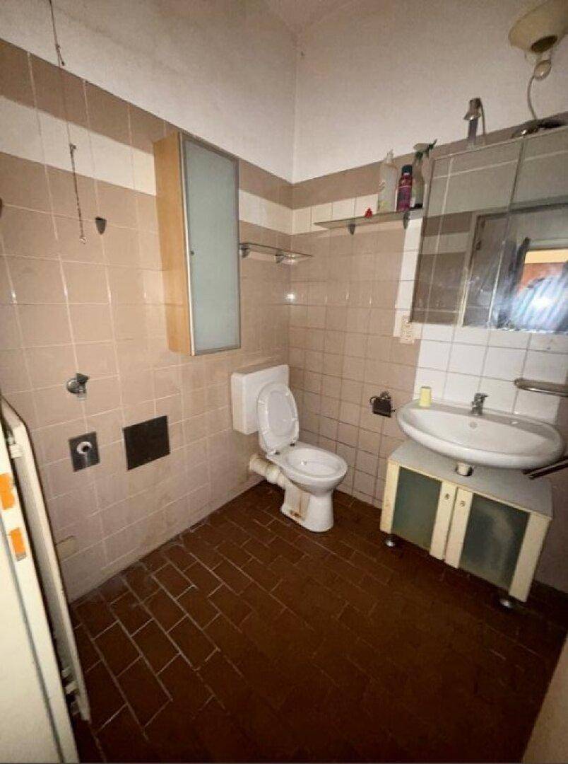 Badezimmer mit beigefarbenen Fliesen, Toilette und Waschbecken, ausgestattet mit Spiegelschrank und Ablagen.