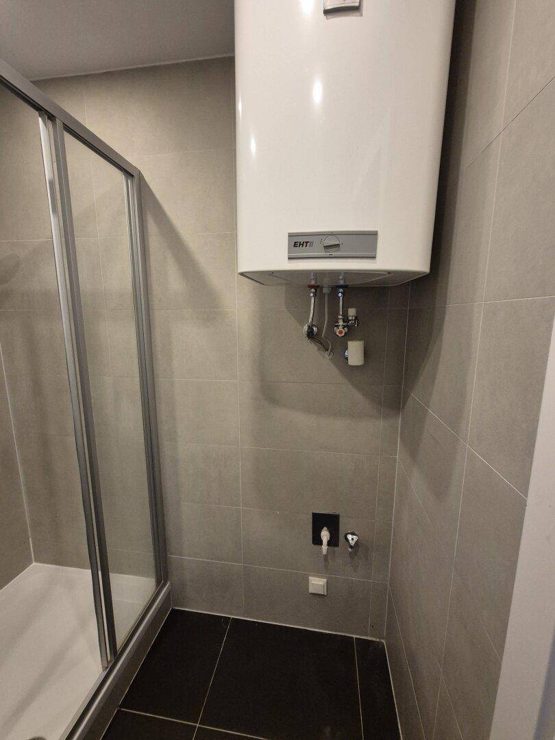 Modernes Badezimmer mit grauen Fliesen, Duschkabine und einem Warmwasserboiler.