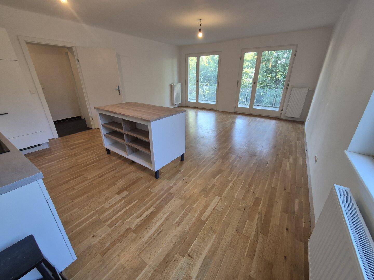 Offener Wohnbereich mit Holzboden, Kücheninsel und großen Fenstertüren zum Außenbereich.