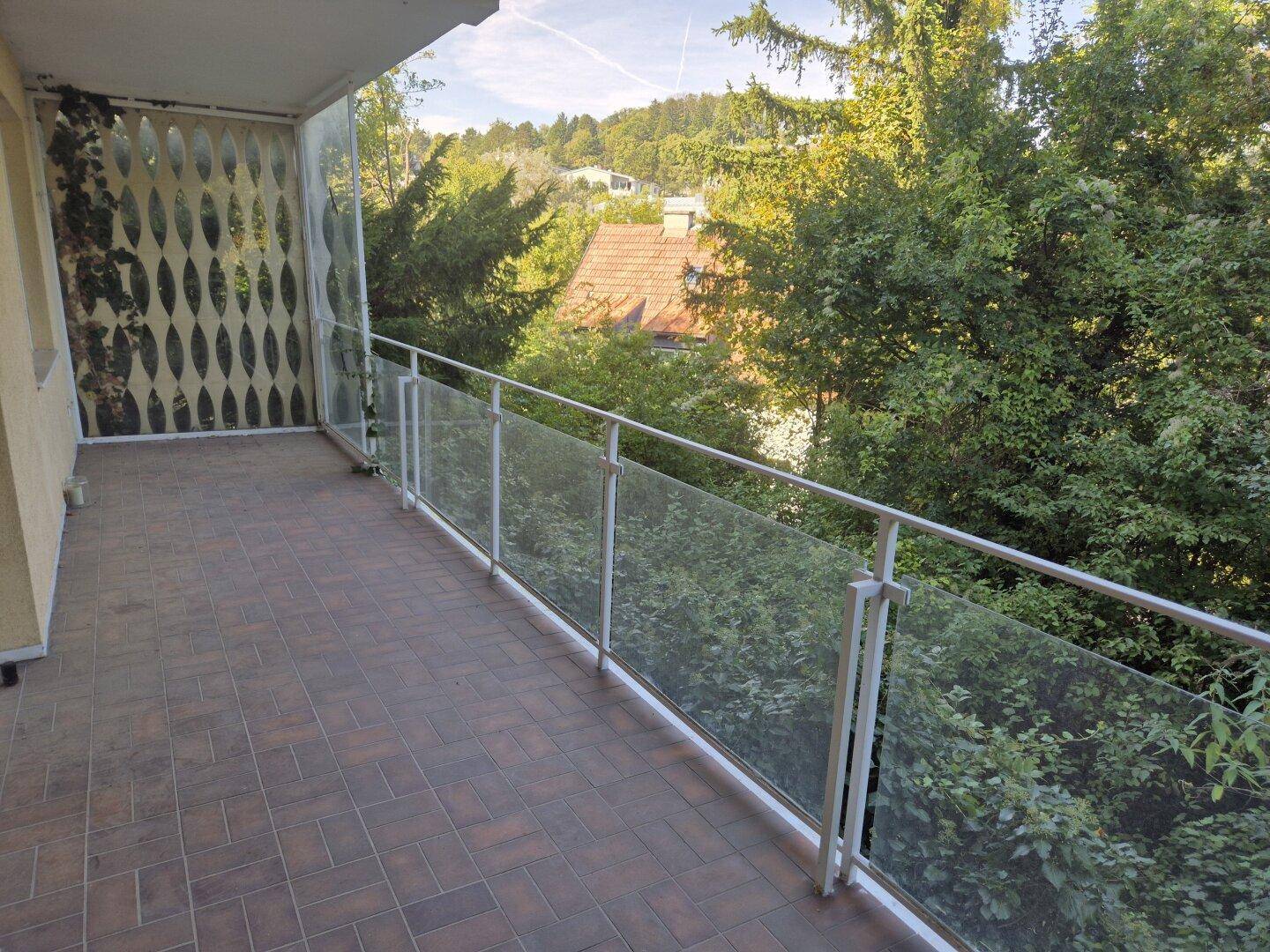 Heller Balkon mit modernem Glasgeländer und weitem Blick über grüne Hügel und Dächer.