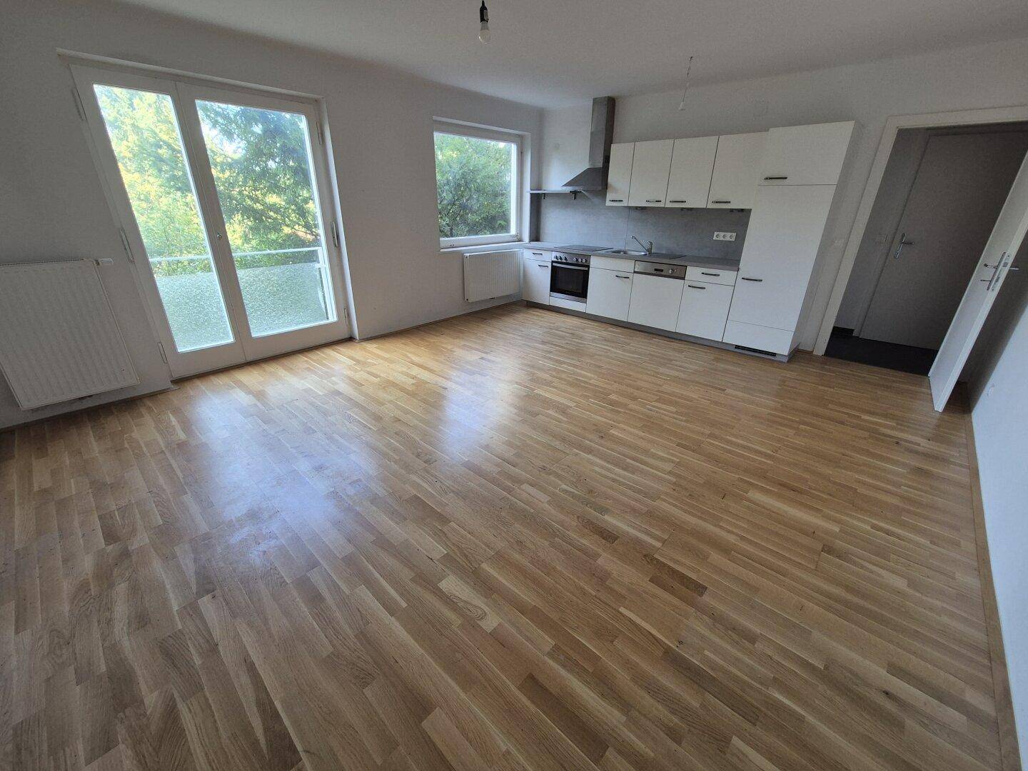 Geräumiger Wohnbereich mit offener Küche, Parkettboden und Zugang zum Balkon.