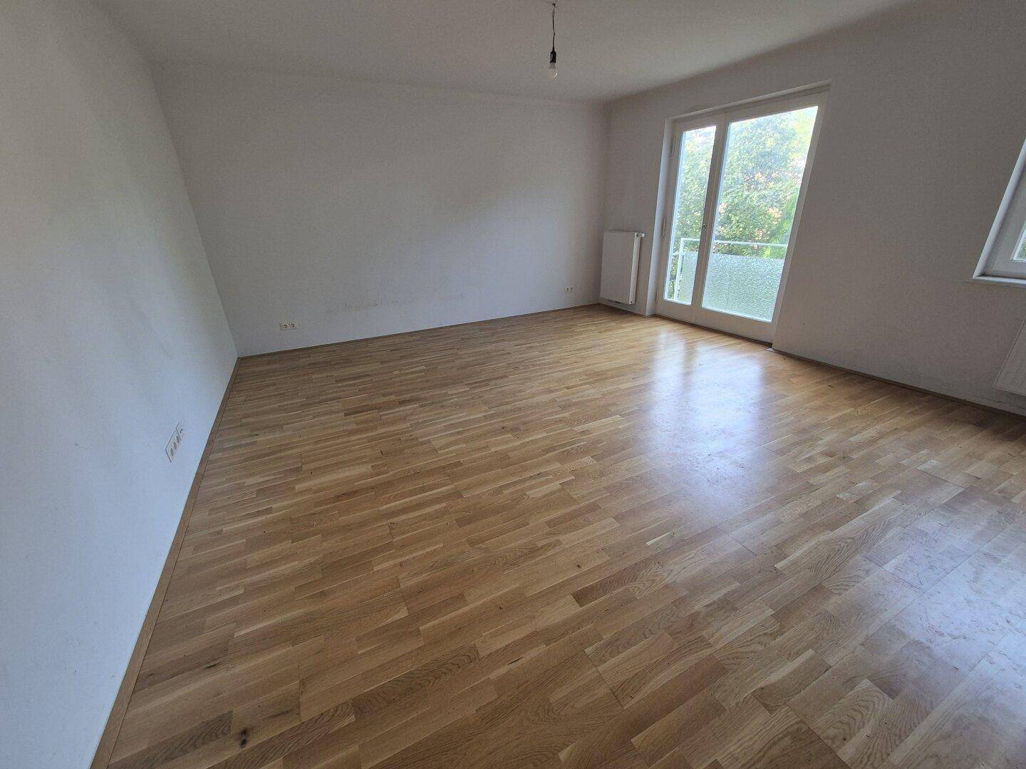 Heller Wohnraum mit Parkettboden und großem Fenster, das viel Tageslicht hereinlässt.