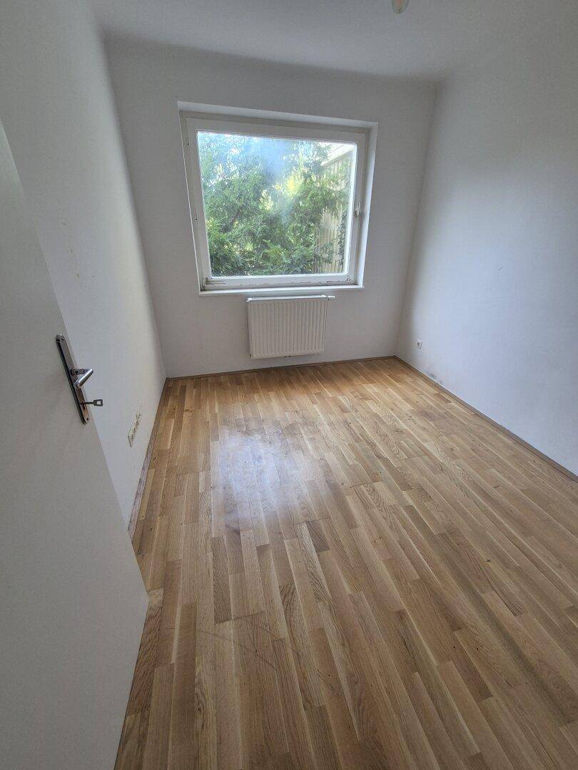 Helles Zimmer mit Parkettboden und Fenster zum grünen Außenbereich, ideal als Schlafzimmer.