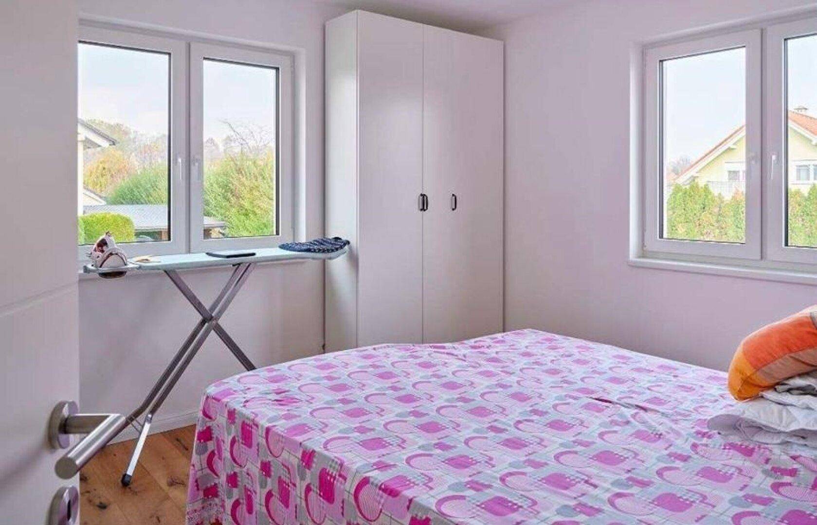 Gemütliches Schlafzimmer mit zwei Fenstern, Holzboden und Kleiderschrank.