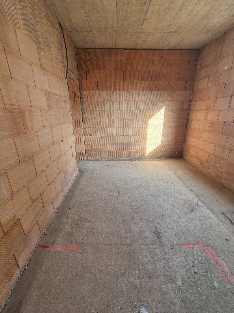 Leeres Rohbauzimmer mit Ziegelwänden und Betonboden, beleuchtet von Sonnenlicht.