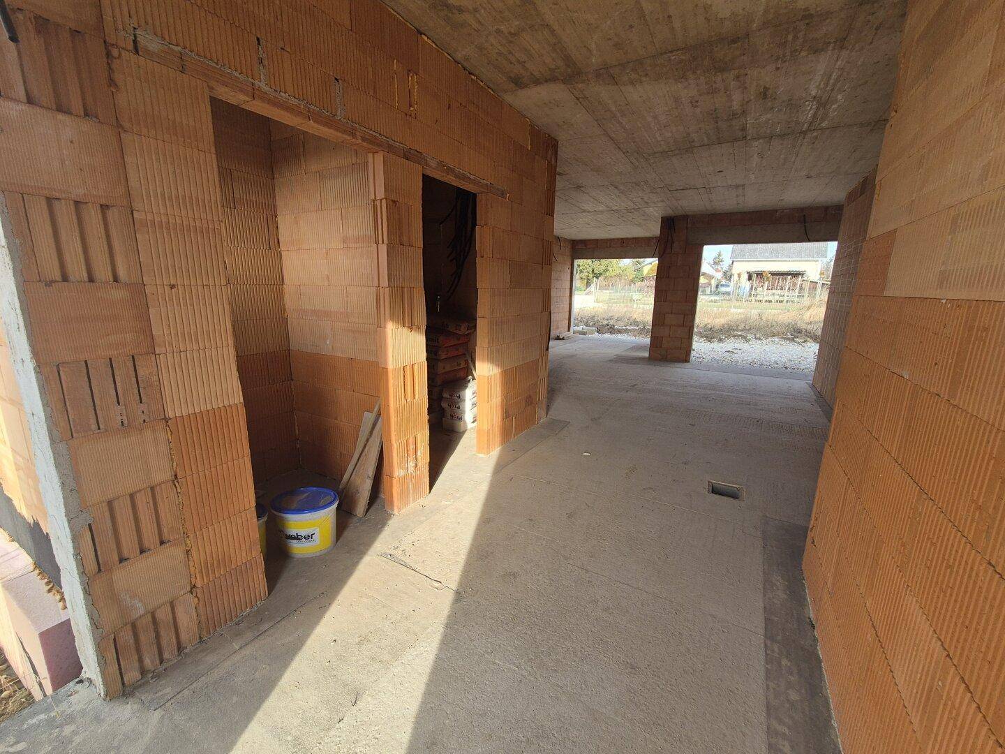 Innenbereich im Rohbau mit Ziegelwänden, Betonboden und Zugang zu weiteren Räumen.