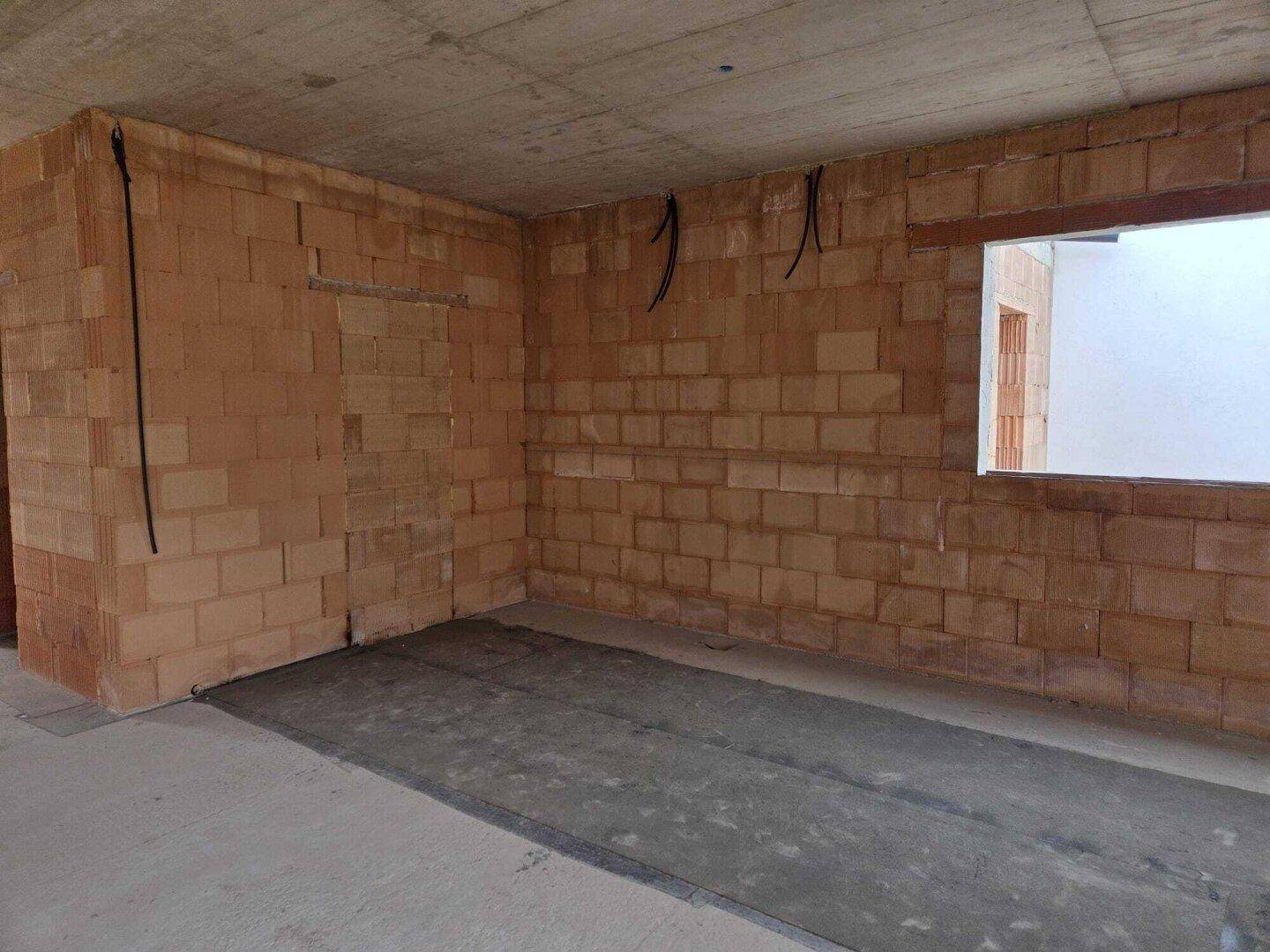 Innenraum im Rohbau mit Ziegelwänden, Betonboden und Fensteröffnung, bereit für den Ausbau.