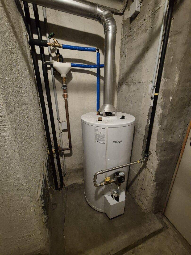 Warmwasserbereiter in einem Technikraum mit sichtbaren Rohren und Betonwänden.