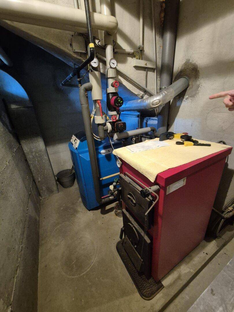 Heizungsanlage mit Kessel und Holzofen in einem Kellerraum mit Betonwänden.