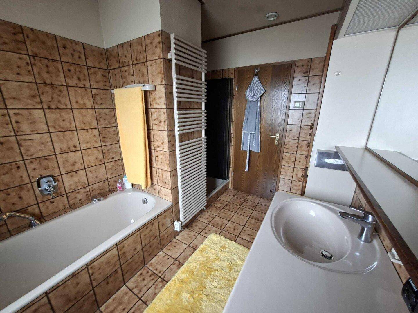 Badezimmer mit Badewanne, Waschbecken und braunen Fliesen, ausgestattet mit Handtuchheizkörper.