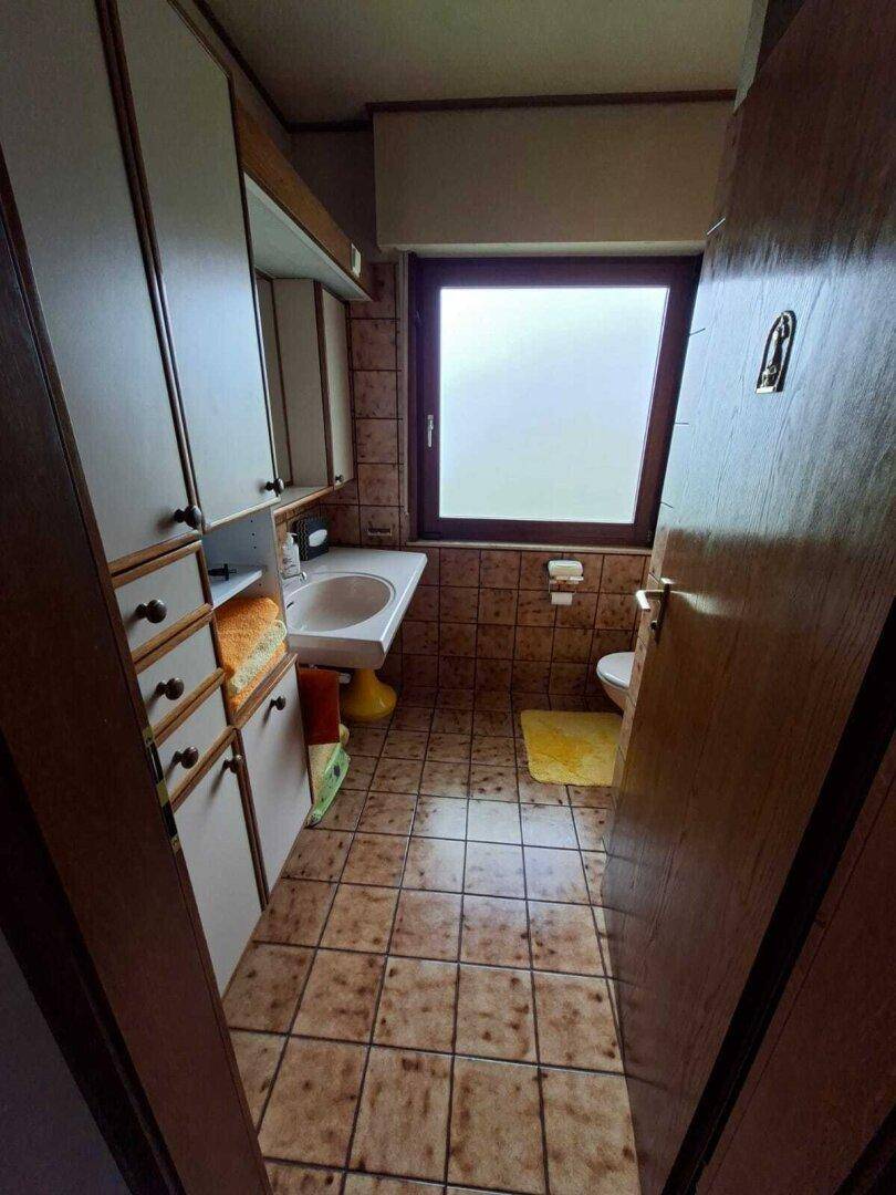 Badezimmer mit Waschbecken, Toilette und Fenster, umgeben von braunen Wand- und Bodenfliesen.