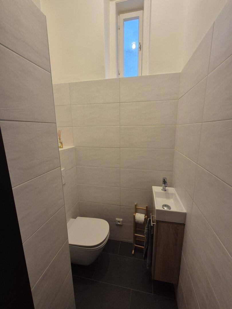 Kleines Gäste-WC mit Wand-WC, kleinem Waschbecken und Fenster auf dunklem Fliesenboden.