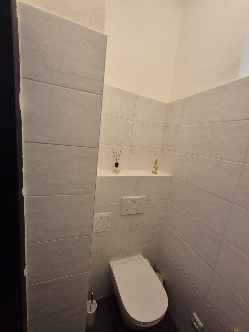 Separate Toilette mit Wand-WC und integrierter Ablagefläche in einem hell gefliesten Raum.