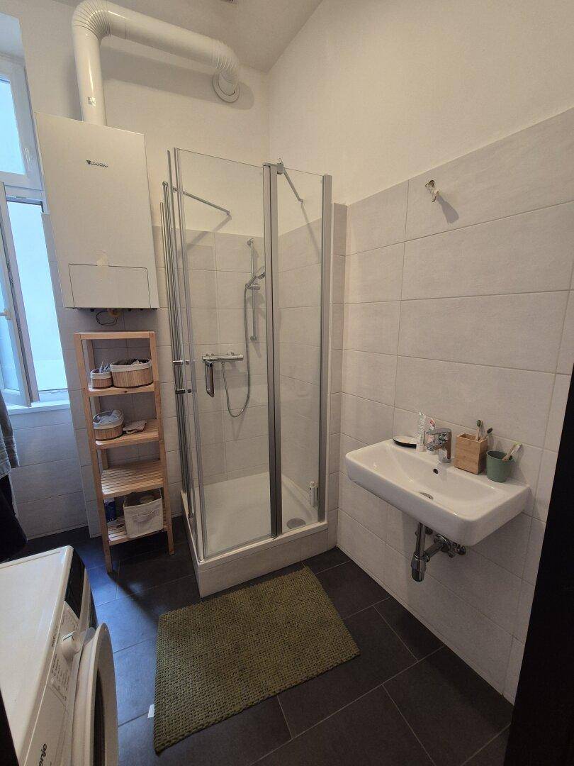 Badezimmer mit Duschkabine, Waschbecken und Waschmaschine auf dunklen Bodenfliesen.
