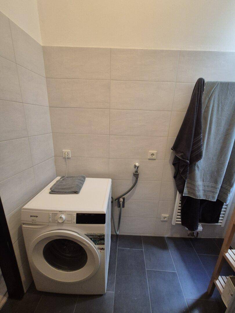Waschmaschine in einem Badezimmer mit hellen Wandfliesen und dunklem Fliesenboden.