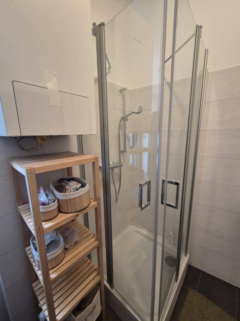 Duschkabine mit Glaswänden in einem hell gefliesten Badezimmer, daneben ein Holzregal.