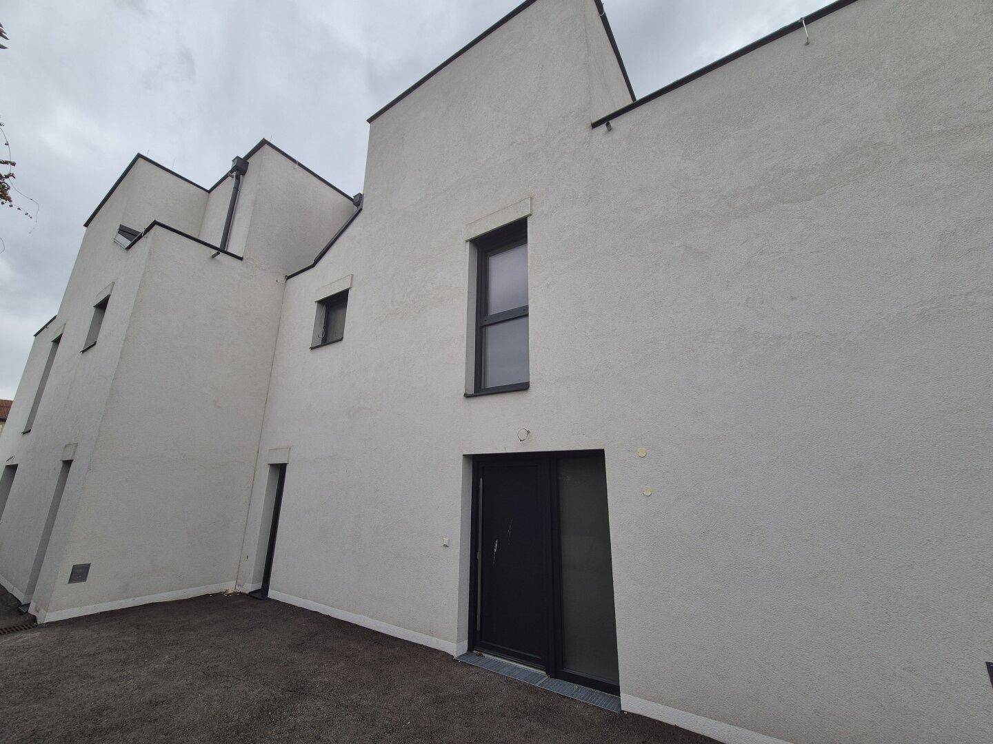 Moderne Gebäudefassade mit Eingangstür und verschiedenen Fenstergrößen unter bewölktem Himmel.
