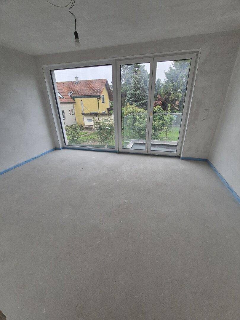 Unfertiger Innenraum mit großem Fenster und Zugang zu einem Balkon, bereit für den Ausbau.