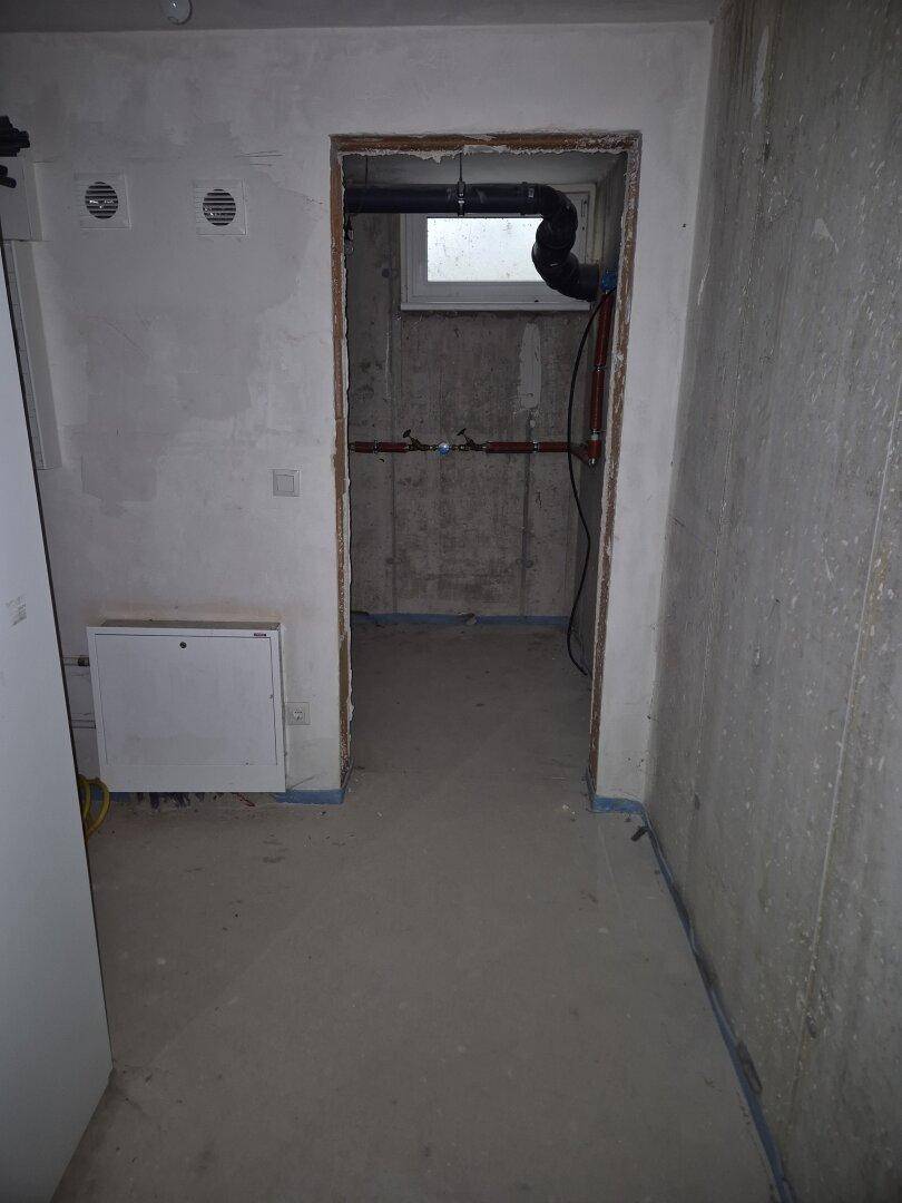 Kellerraum im Rohbau mit kleinem Fenster und sichtbaren Versorgungsleitungen, typisch für einen Neubau.