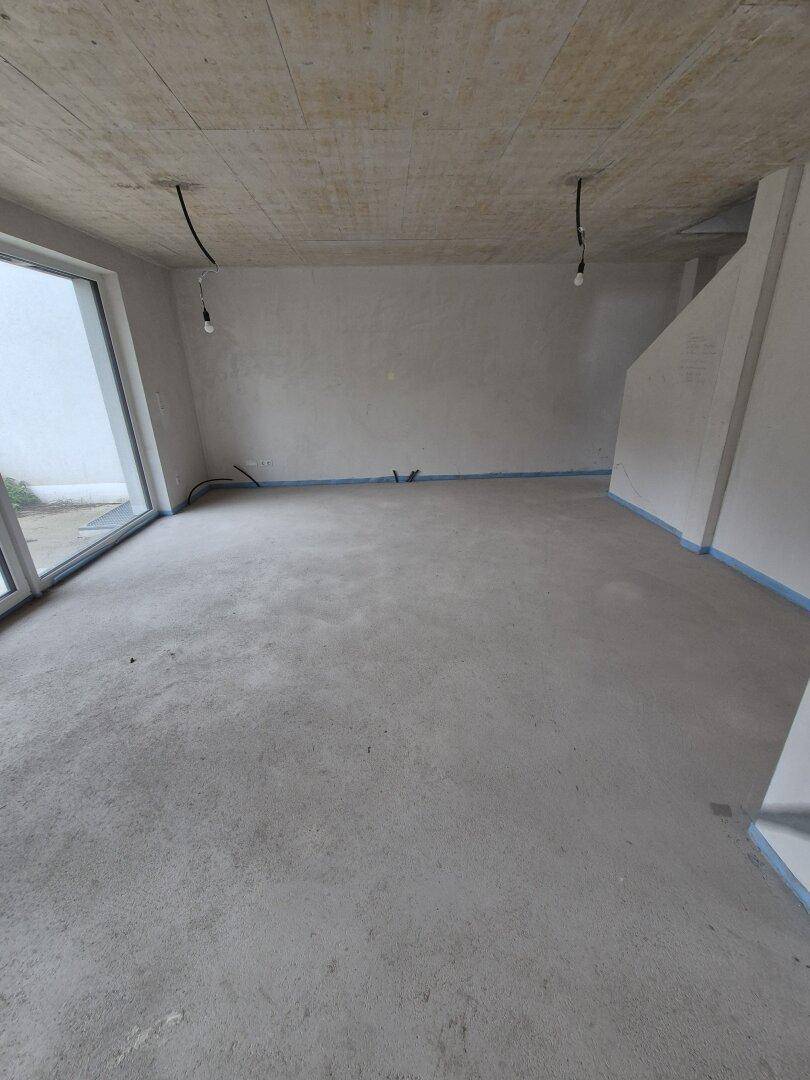 Geräumiger Wohnbereich im Rohbau mit großen Fenstern und Blick nach draußen, viel natürliches Licht.