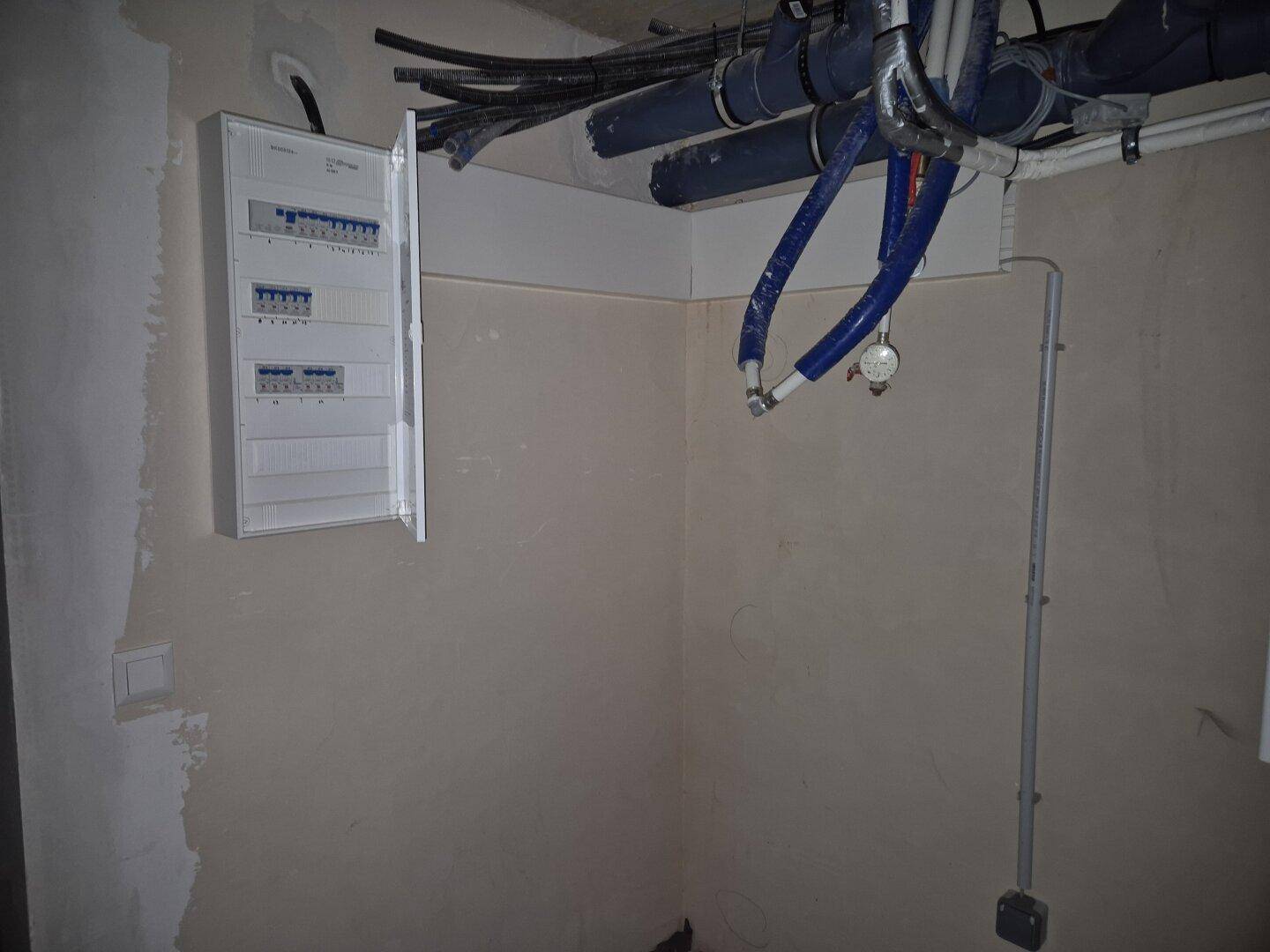 Technikraum mit offenliegenden Rohren und einem Elektroverteilerkasten an der Wand im Rohbau.