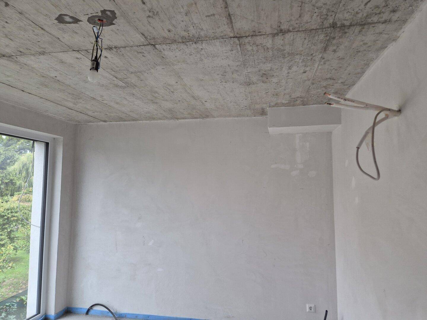 Unfertiger Innenraum mit Betonrohdecke und großem Fenster, bereit für den Ausbau.