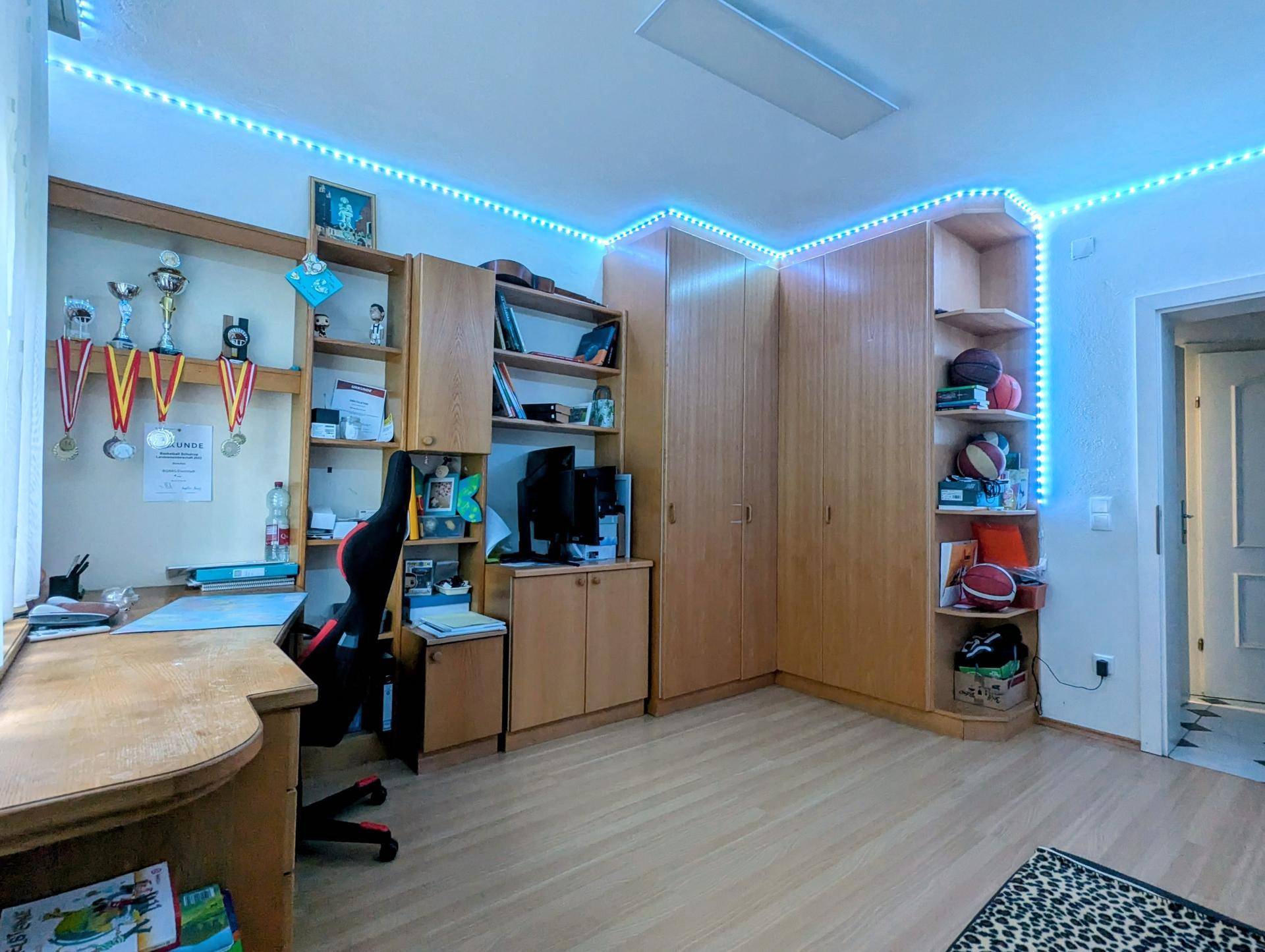 Jugendzimmer mit Holzmöbeln, Regalsystemen und blauer LED-Streifenbeleuchtung.