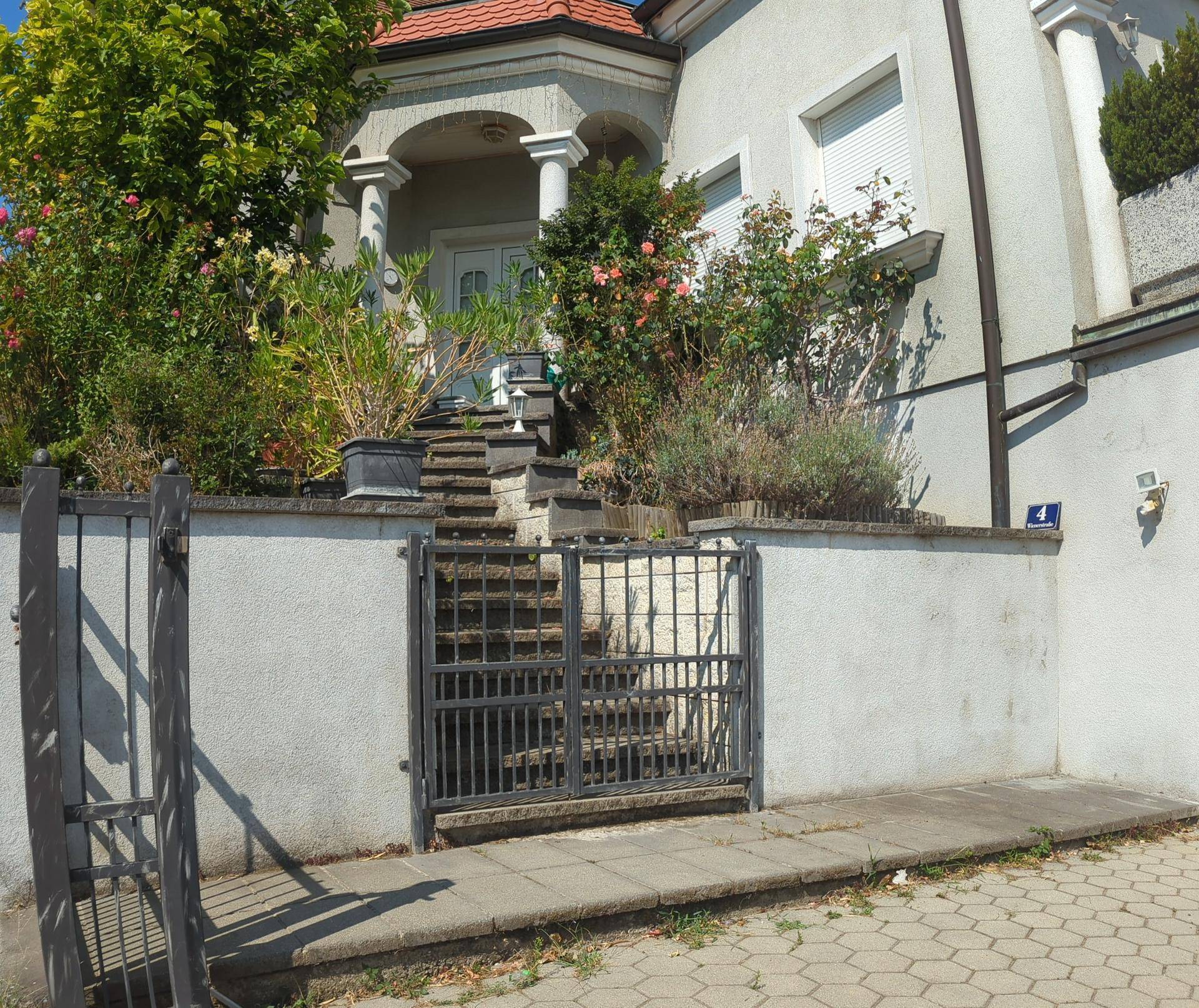 Eingangsbereich des Hauses mit schmiedeeisernem Tor und Steintreppe, umgeben von Pflanzen.