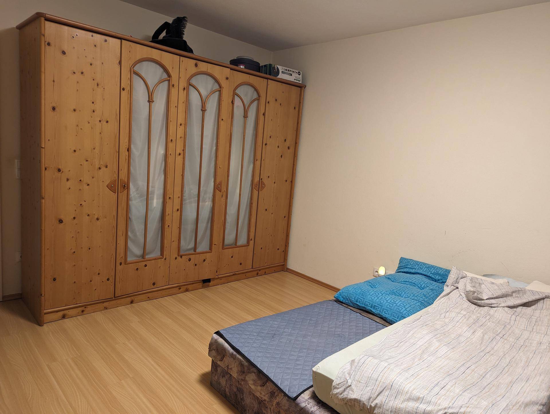Schlafzimmer mit großem Holzschrank und einfacher Bettausstattung auf hellem Boden.