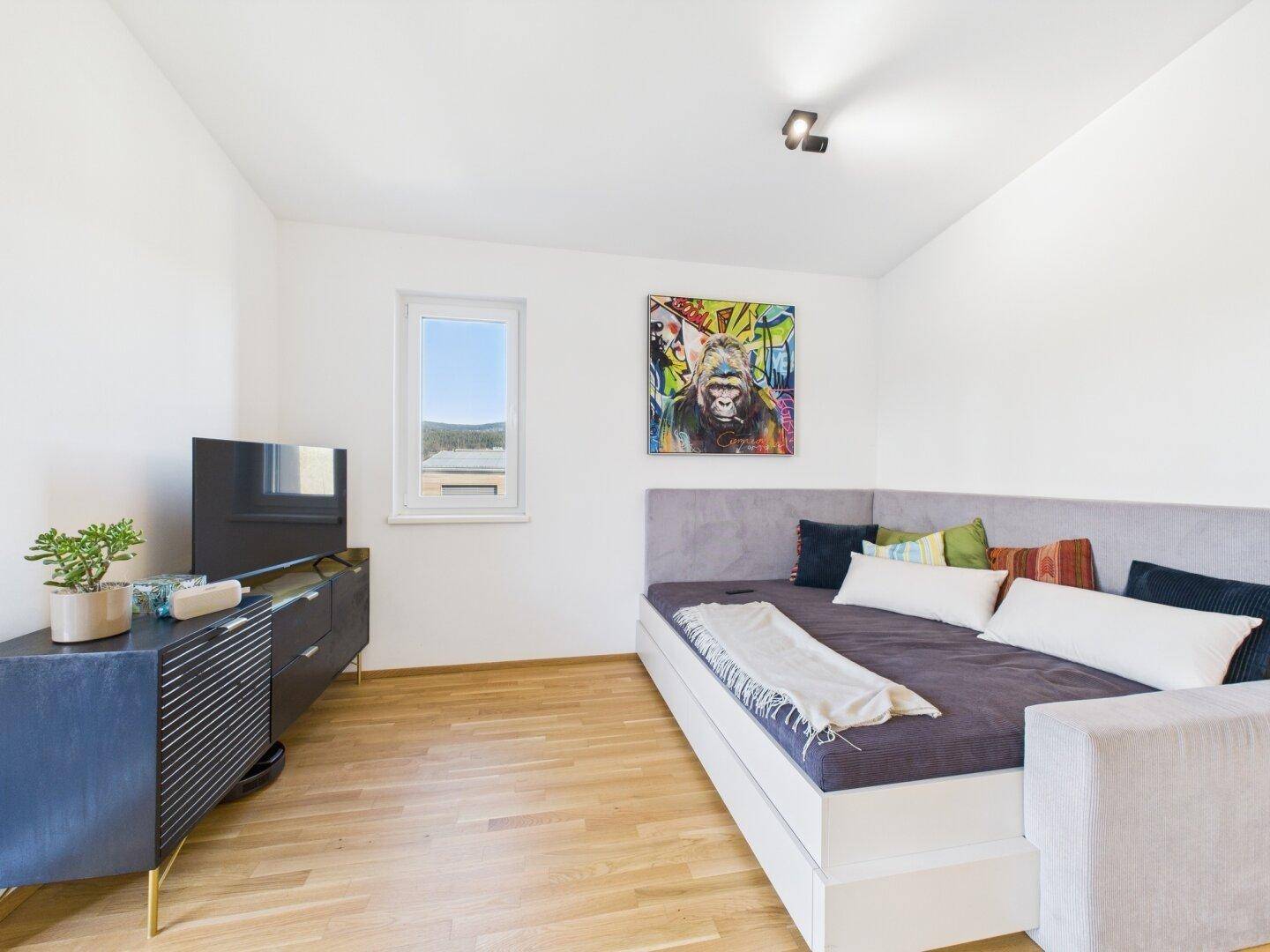 Helles Schlafzimmer mit Tagesbett, Fernseher und Fenster mit grünem Ausblick.