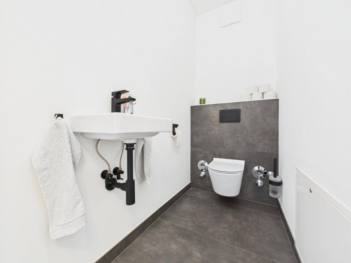 Modernes Gäste-WC mit Wand-WC, kleinem Waschbecken und dunklen Fliesen.
