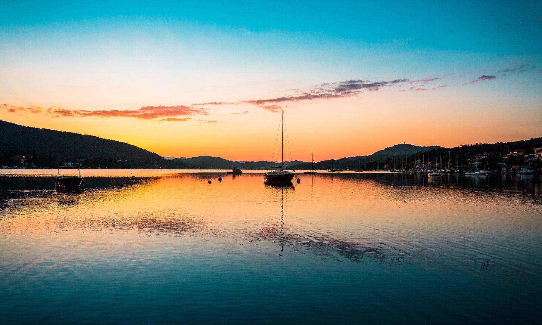 Stimmungsvoller Sonnenuntergang über dem See mit Segelbooten und einer malerischen Bergkulisse.