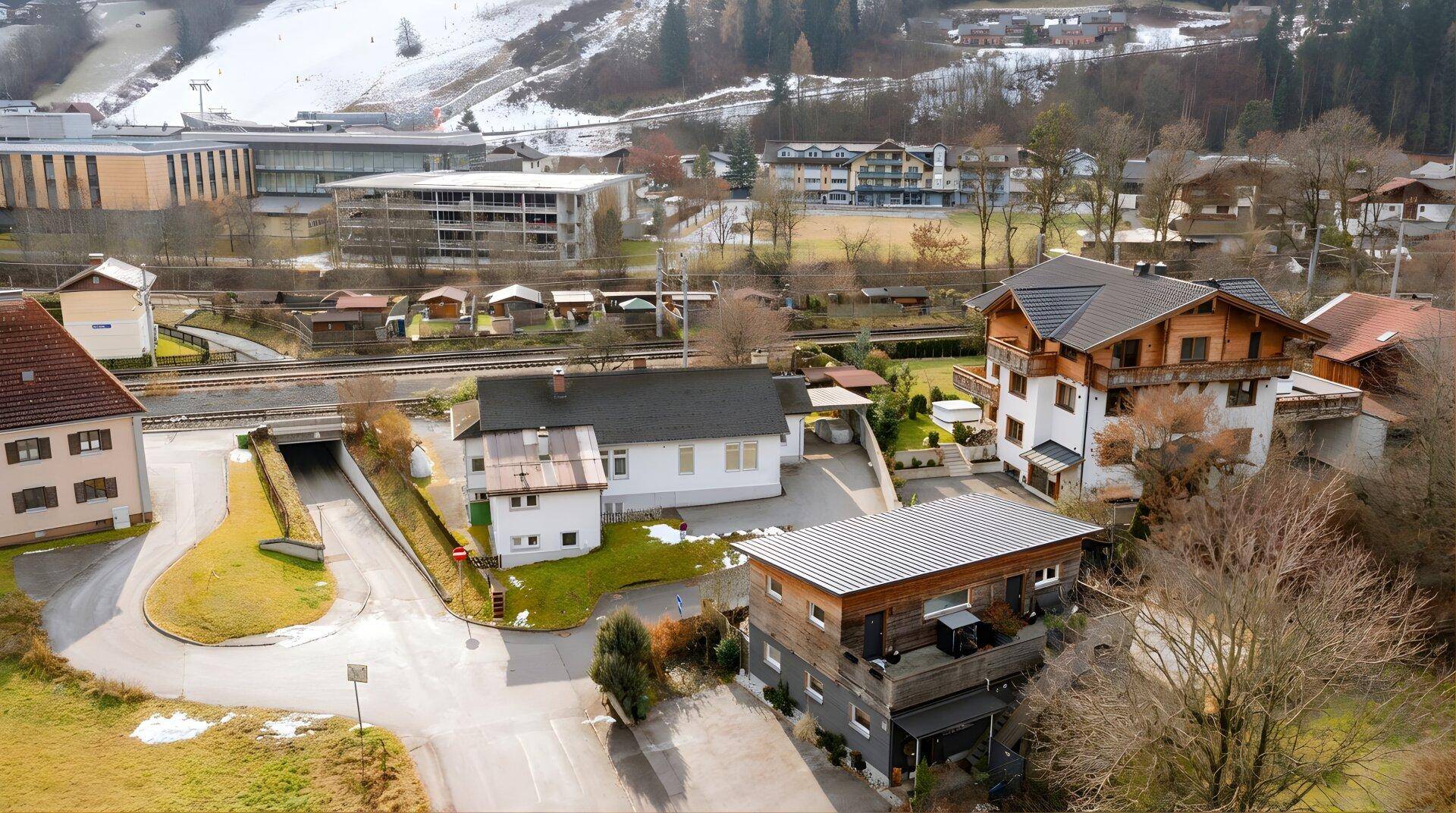 Luftaufnahme einer ländlichen Siedlung mit Häusern und schneebedeckten Bergen im Hintergrund.