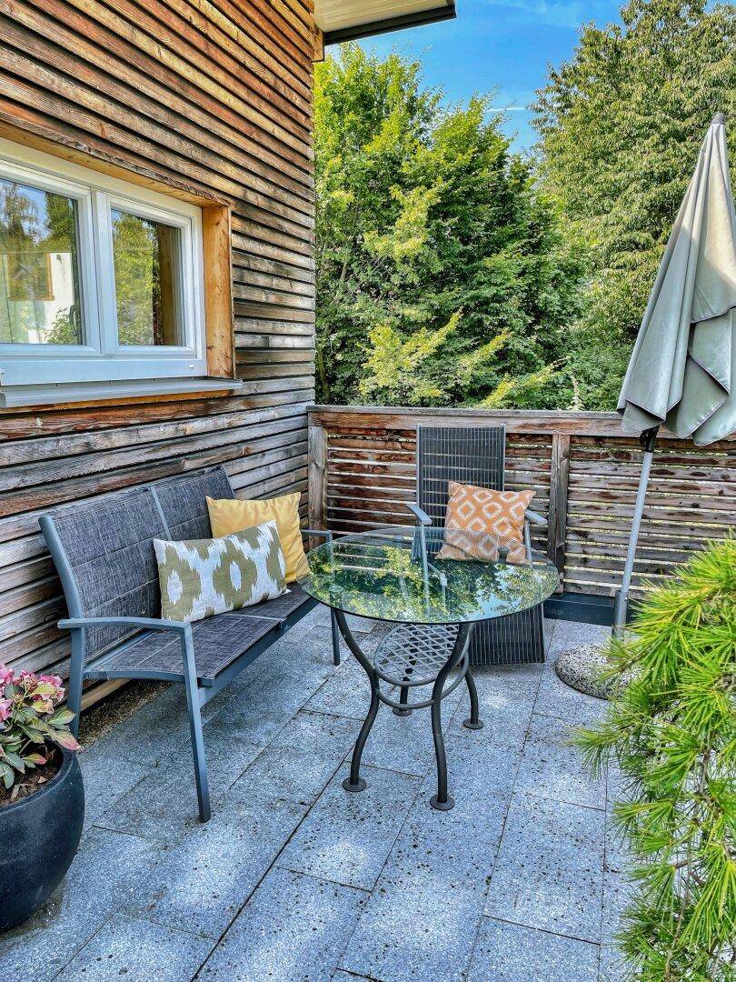 Kleine Terrasse mit Sitzgelegenheiten, Glastisch und Holzwand.
