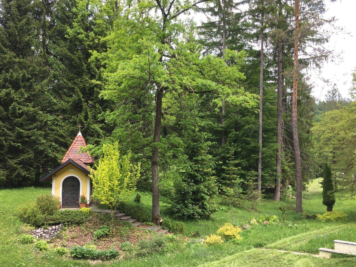 Kleine, charmante Kapelle in einem grünen Gartenbereich, umgeben von hohen Bäumen und Sträuchern.