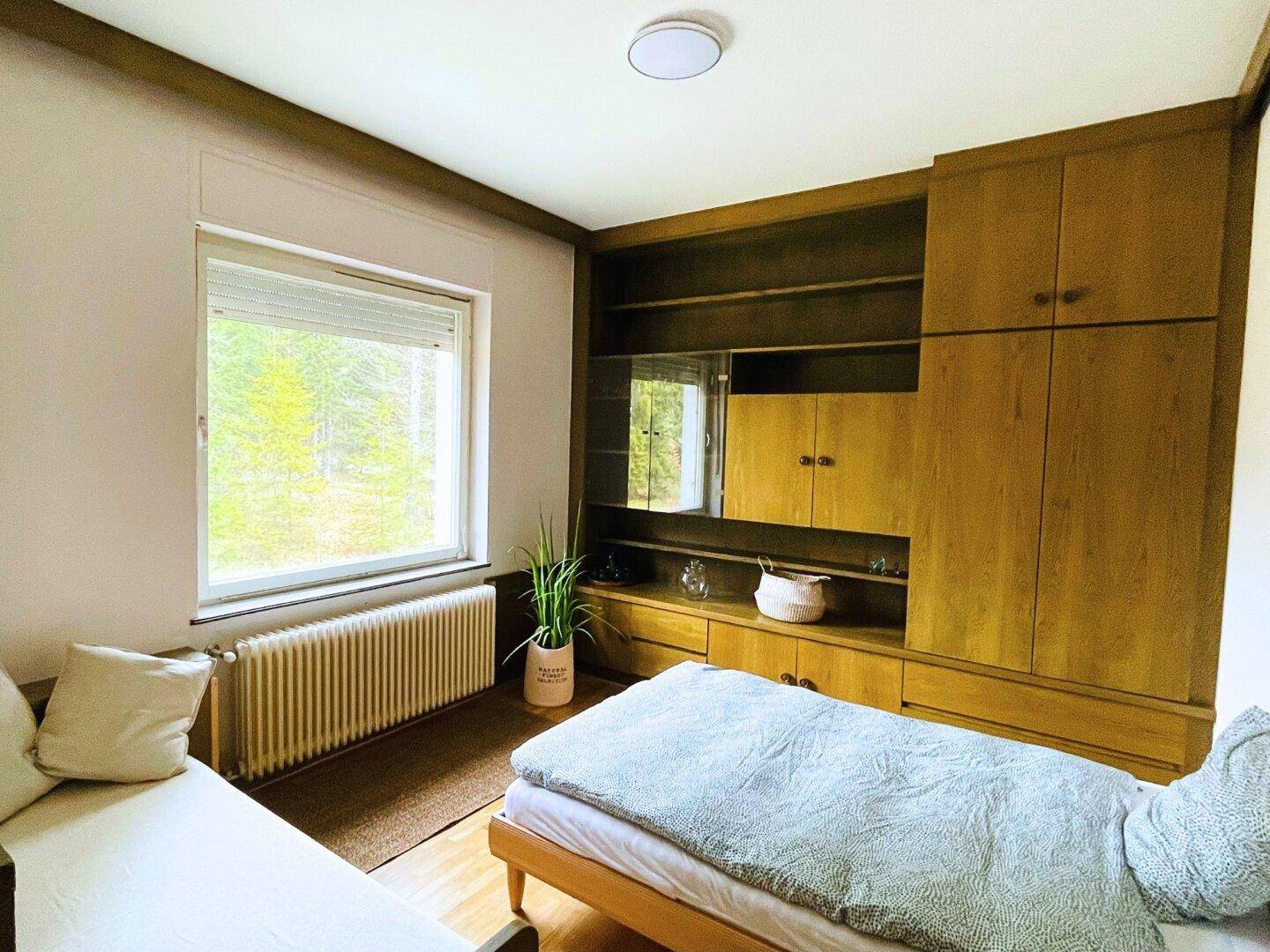 Schlafzimmer mit Einzelbett, Holzmöbeln und einem großen Fenster mit Blick ins Grüne.
