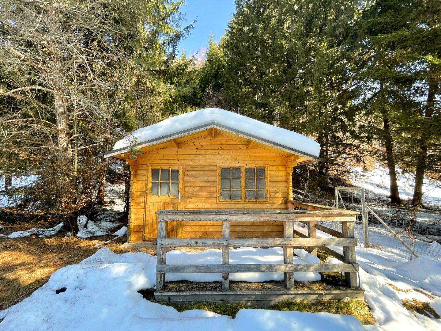 Kleine Holzhütte mit Schnee auf dem Dach, umgeben von verschneiten Bäumen im Winter.