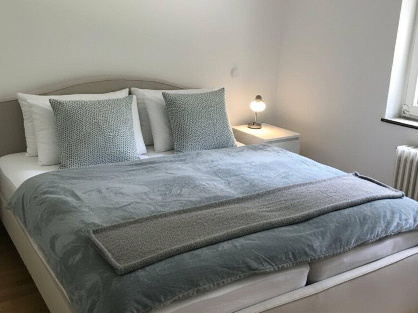 Helles Schlafzimmer mit Doppelbett, Nachttisch und Fenster, das viel Tageslicht hereinlässt.