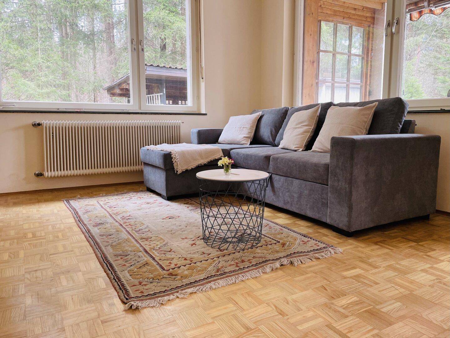 Gemütliches Wohnzimmer mit grauem Sofa, Parkettboden und Blick ins Grüne durch große Fenster.