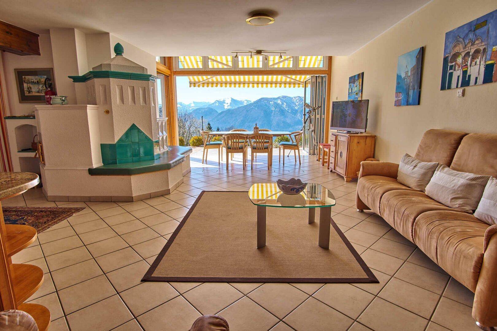 Wohn-Essbereich mit traditionellem Kachelofen, Fliesenboden und Zugang zur Terrasse mit Bergblick.